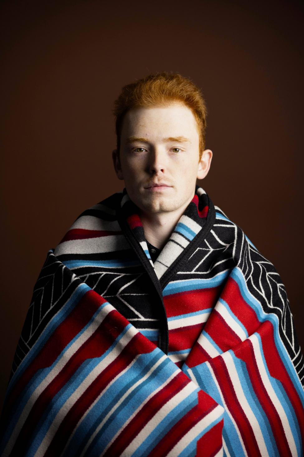 Studio portrait of a person with short red hair wrapped in a patterned blanket featuring red, blue, black, and white stripes and geometric designs. The person faces the camera against a dark brown background, with the blanket draped around the shoulders and covering most of the body.