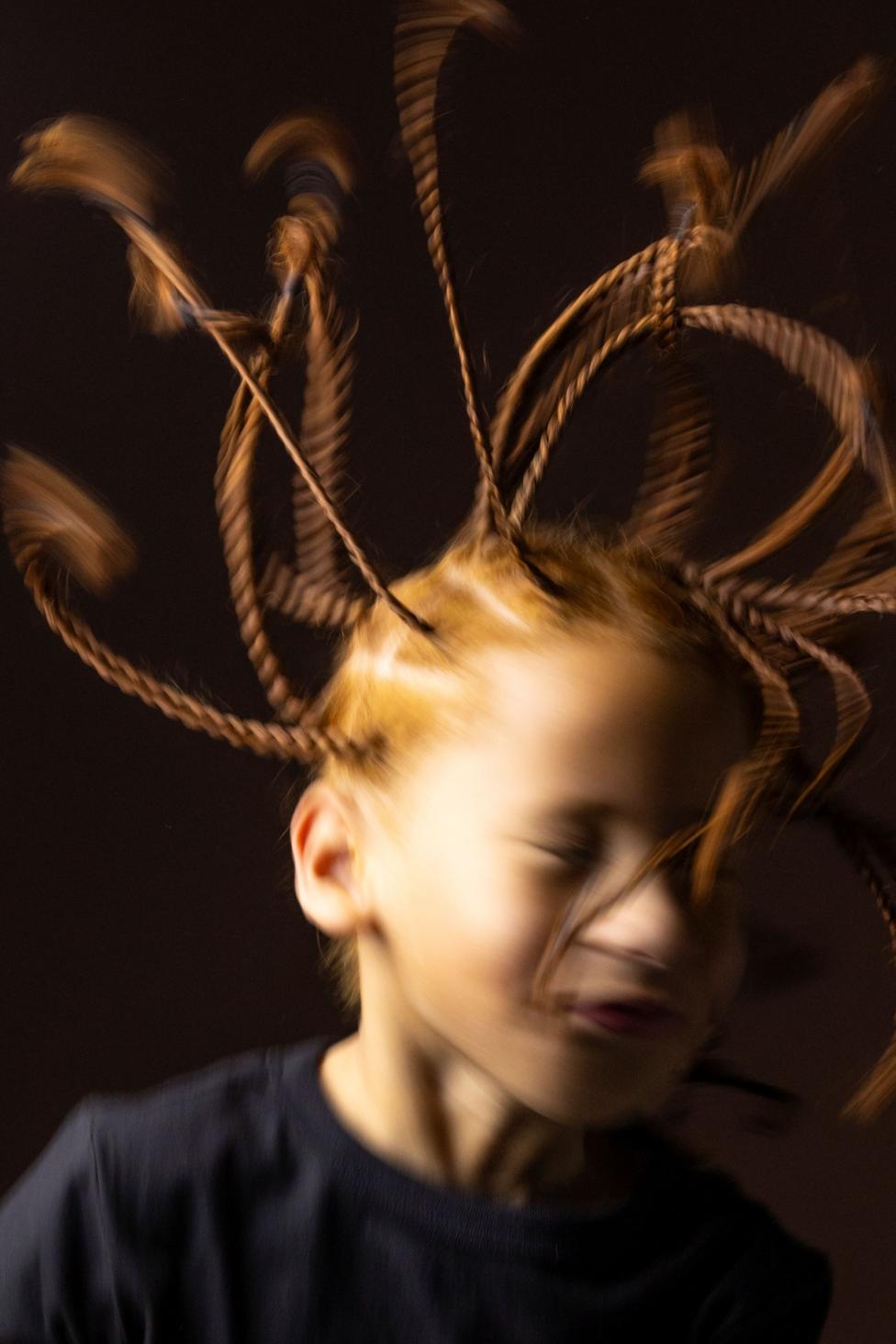 Artistic close-up portrait of a child with multiple braided strands of hair extended outward in motion, creating a dynamic blurred effect. The child wears a dark top and is photographed against a dark brown background with warm studio lighting that highlights the movement and texture of the braids.