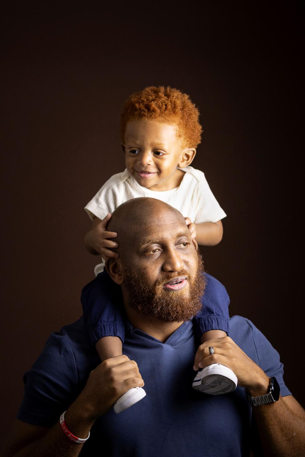 Studio portrait of an adult wearing a blue shirt with a young child seated on the adult’s shoulders. The child wears a light-colored T-shirt, dark trousers, and white shoes, with hands resting on the adult’s head. Both are photographed against a dark brown background with warm, soft lighting.