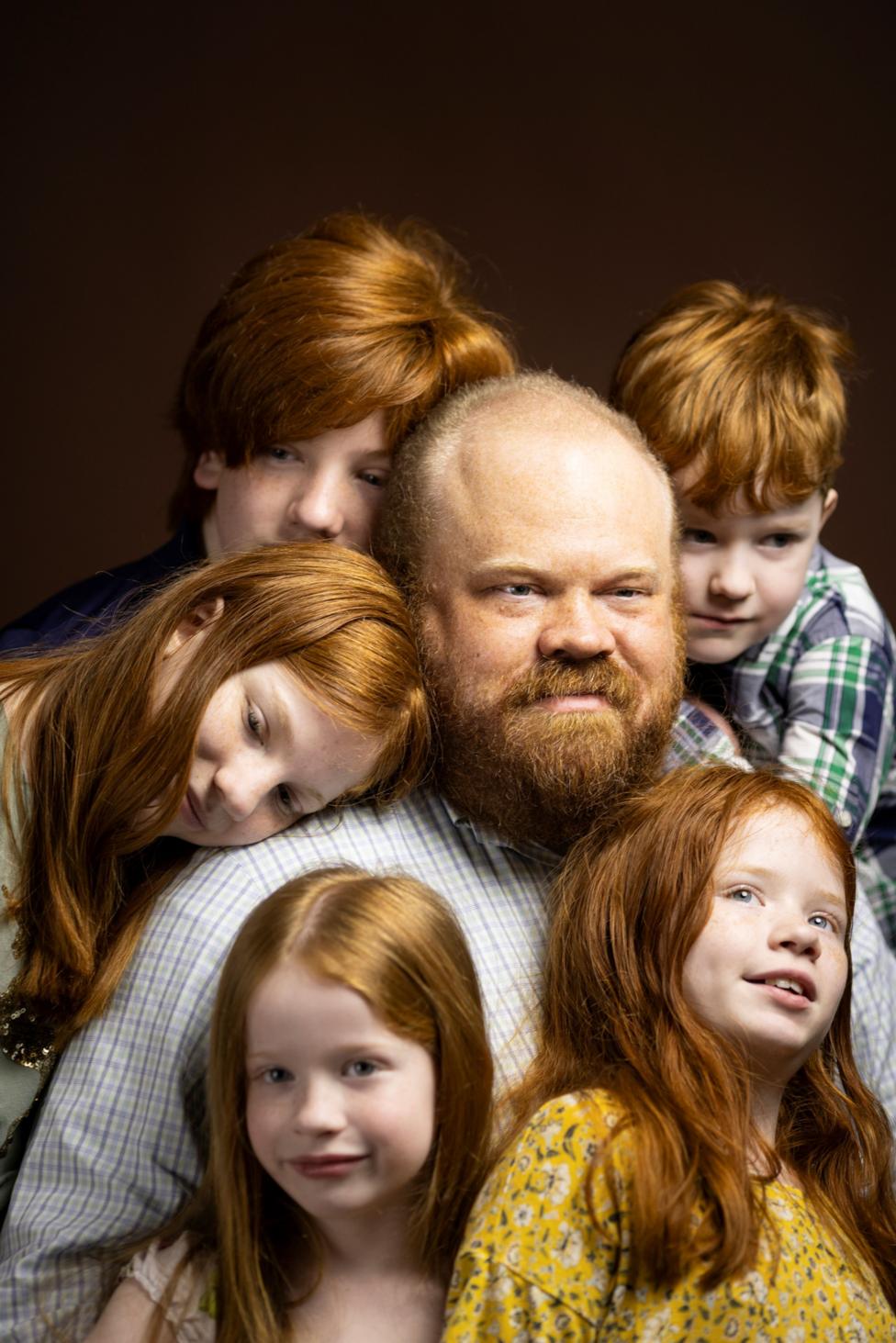 Studio portrait of an adult with a beard seated in the center, surrounded by five children with red hair. Two children lean against the adult’s shoulders, one stands behind, and two are positioned in the foreground. The group is closely gathered against a dark brown background, with warm lighting highlighting their hair and patterned clothing.