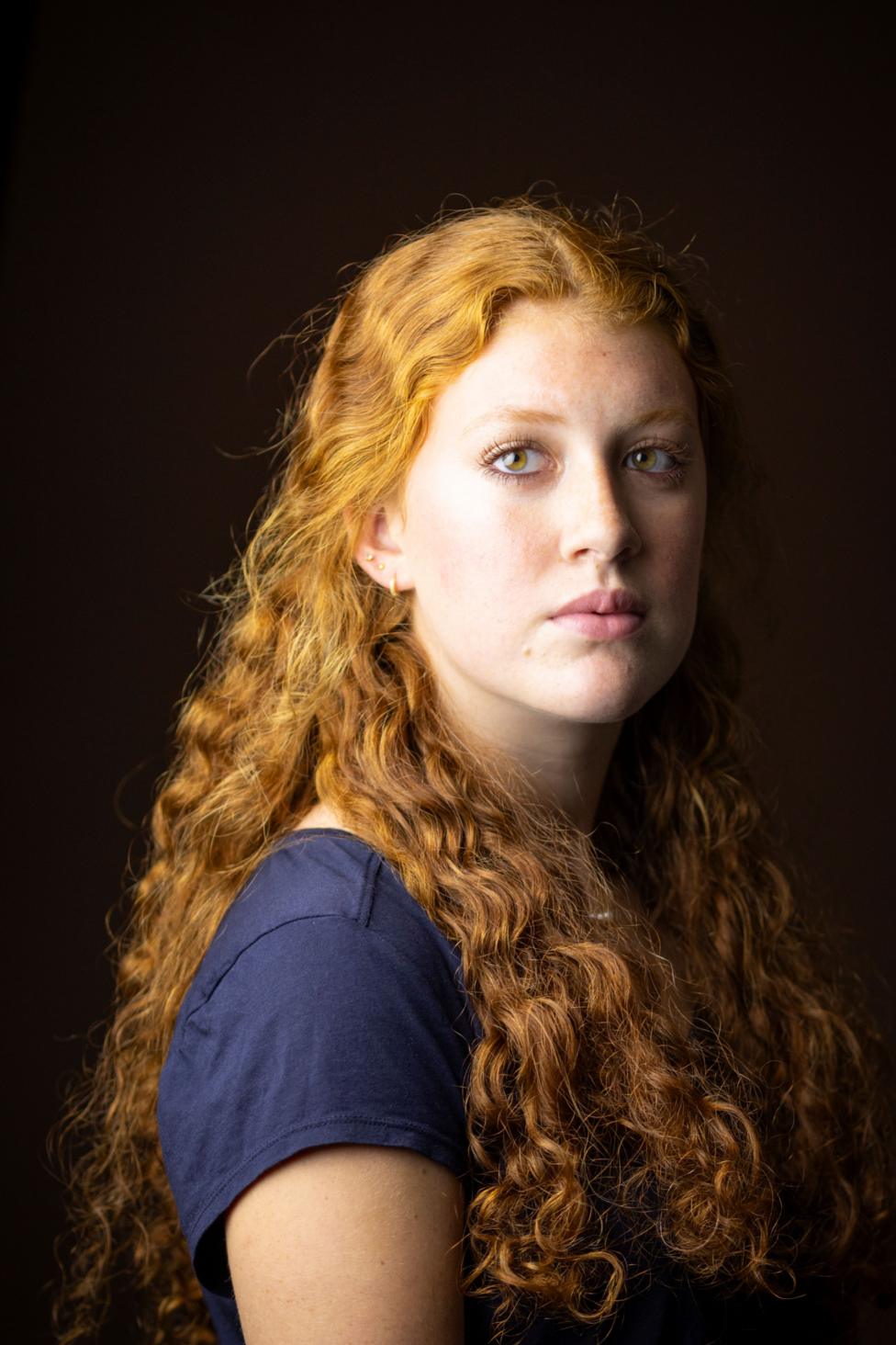 Close-up studio portrait of a person with long, curly red hair cascading over the shoulders, wearing a dark short-sleeved top. The person is turned slightly to one side while looking toward the camera. Soft, warm lighting highlights the texture of the hair against a dark brown background.
