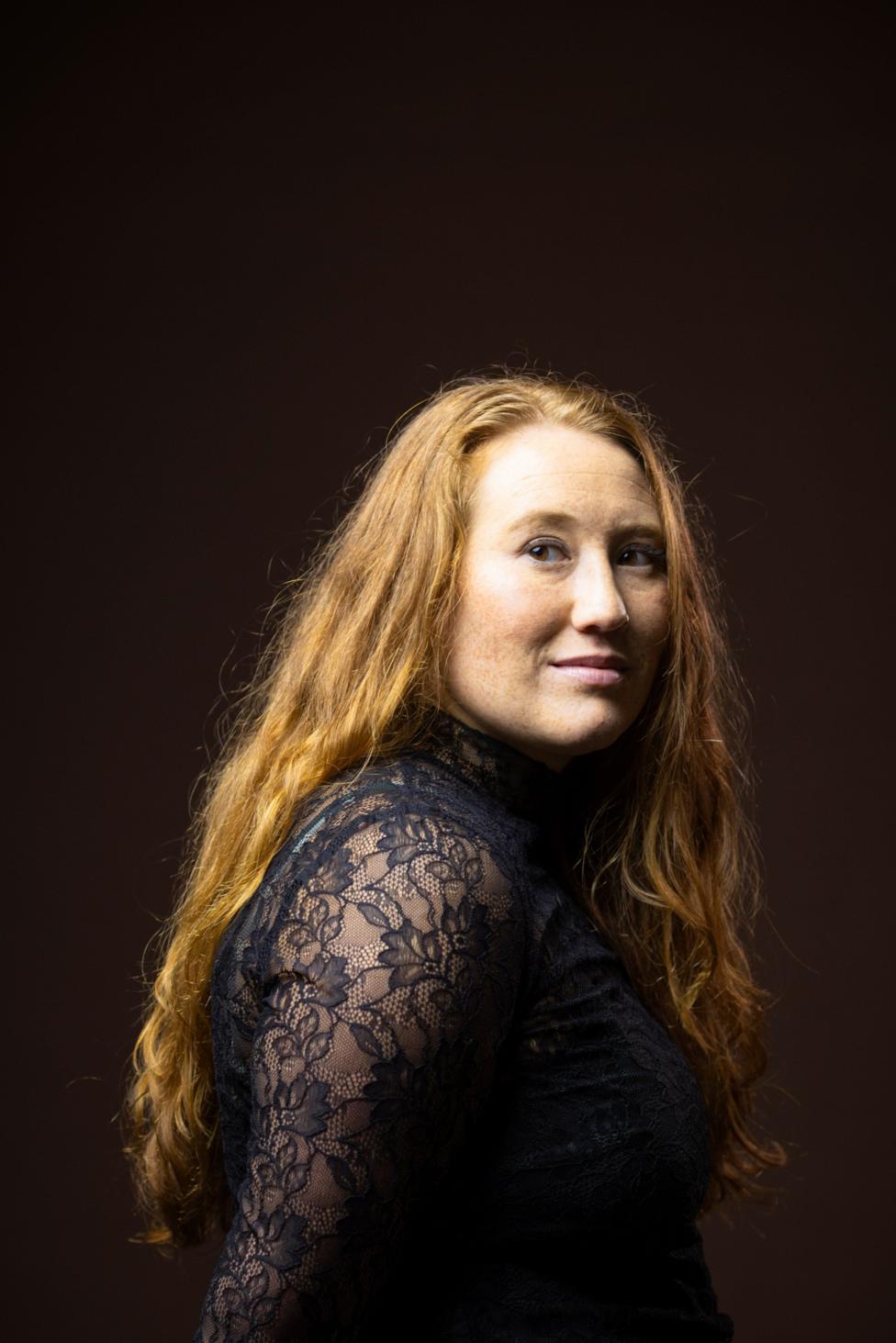 Studio portrait of a person with long, wavy hair wearing a black high-neck lace top with sheer floral-patterned sleeves. The person is turned slightly sideways while looking toward the camera. Warm studio lighting highlights the hair texture and intricate lace details against a dark brown background.