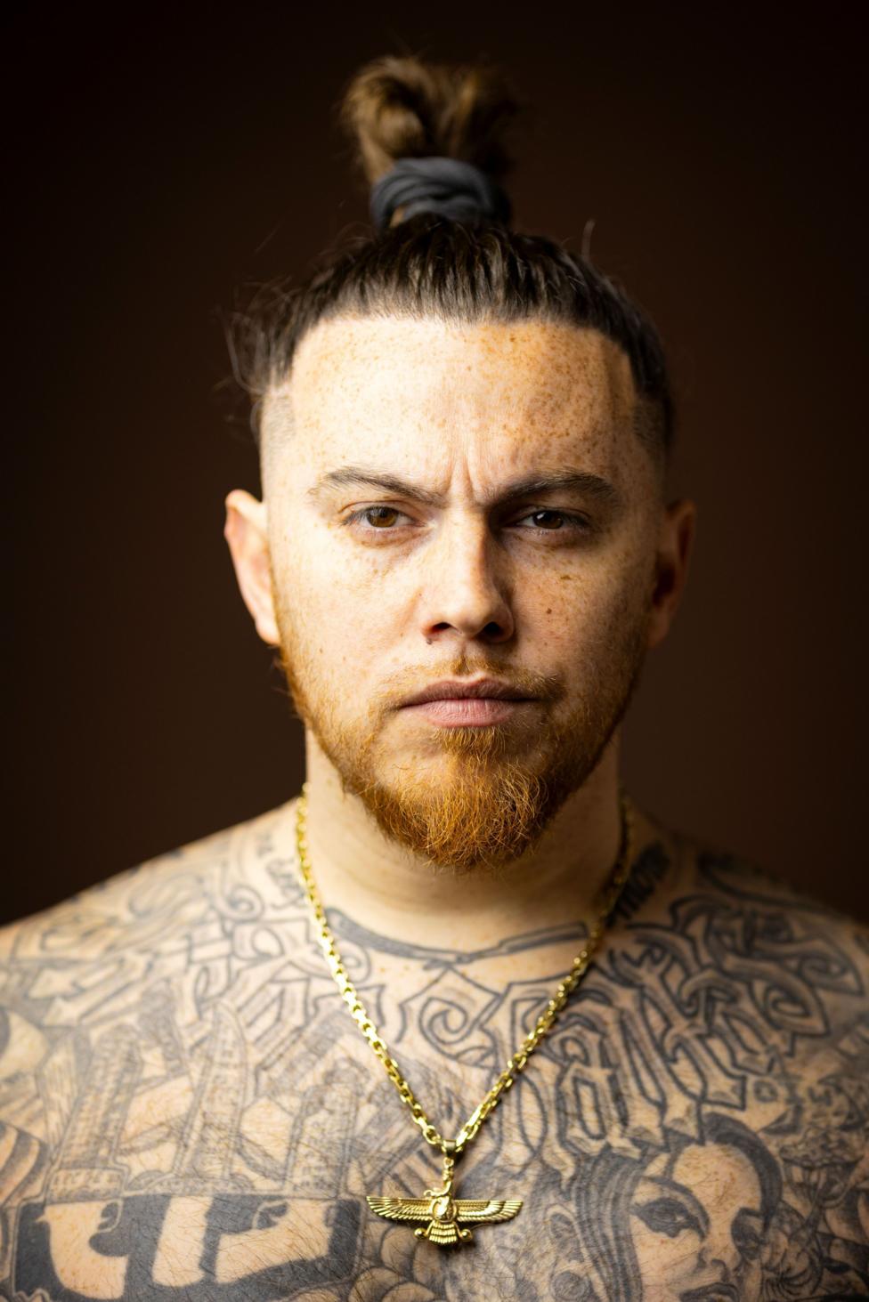 Close-up studio portrait of a person with hair tied in a top knot, freckles, a short beard, and extensive black-and-grey tattoos across the chest and shoulders. The person wears a gold chain necklace with a winged pendant and faces the camera against a dark brown blurred background.