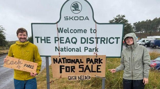 Two people at the side of a road sign which reads SKODA WELCOME TO THE PEAQ DISTRICT NATIONAL PARK. A cardboard sign is hung around the larger sign which reads NATIONAL PARK FOR SALE CASH NEEDED.