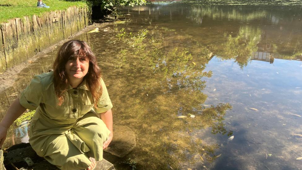 A woman wearing a light green jumpsuit kneeling down by the side of the water. She has wavy brown hair and is smiling while looking up to the camera. She is kneeling next to the river.