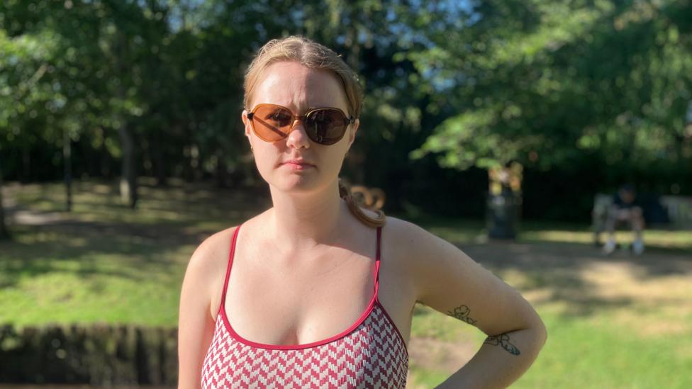 A woman wearing a red tank top with sunglasses while standing outside. Her hair is in a middle parting and braided at the back. She has one hand on her hip and is looking at the camera. Behind her is some grass and trees