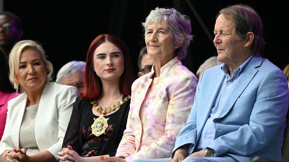 A woman with blonde hair and a cream suit is seen sitting alongside a woman with red hair and a black top who is wearing a mayoral chain. Beside her is a a woman with short grey hair wearing a light floral jacket and a man beside her with brown hair and wearing a cornflower blue shirt and suit. 
