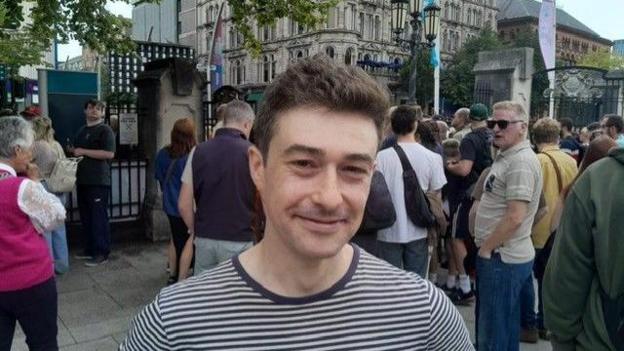 A man wearing a navy and white stripped t shirt is seen smiling at the camera as he stands in front of the city hall in Belfast ahead of the start of the Fleadh Cheoil na hÉireann with several dozen other people standing in the background. 