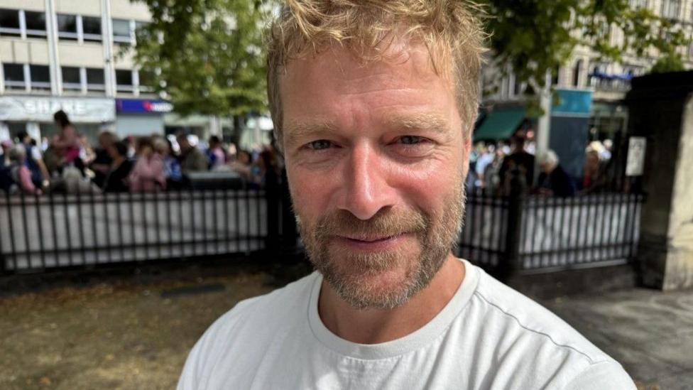 A man with blonde hair and a beard wearing a white t shirt is seen smiling at the camera inside the grounds of Belfast City Hall. Behind him is a black metal fence with crowds gathered behind. 