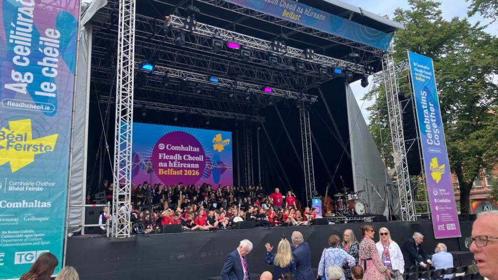 A number of performers are seen seats at the main stage for Fleadh Cheoil na hÉireann outside Belfast City Hall while a number of other people are seen in the foreground. 