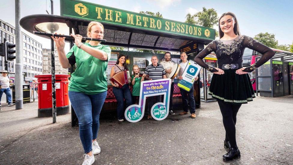 Two women, one holding a flute and wearing blue jeans and a green t-shirt, the other with long brown hair and wearing a black Irish dancing dress. They are standing in front of a stall with a sign above it reading 