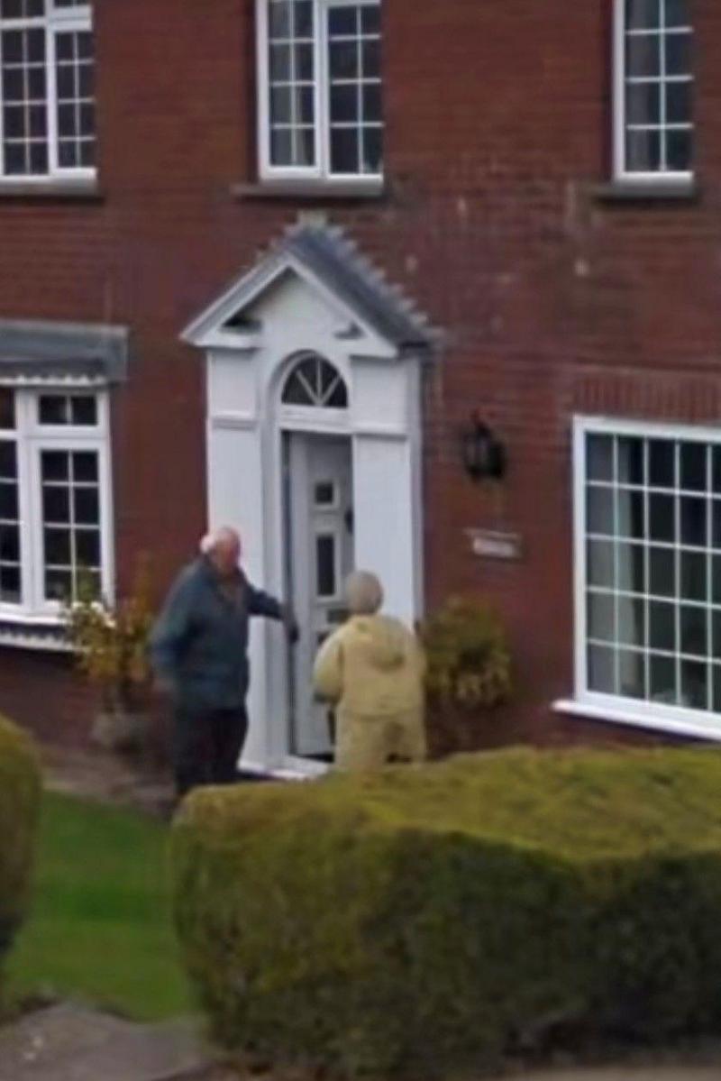 A man and a woman are stood outside a large red-brick home. He is holding the door open for her in the dooway.