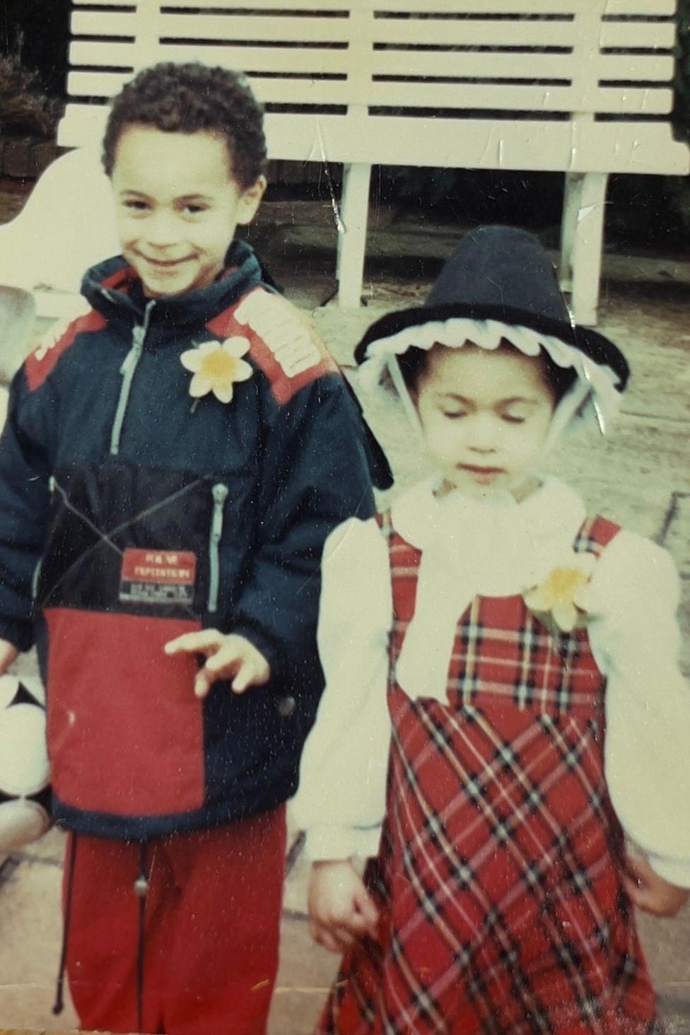 A young girl is wearing a traditional Welsh dress and a daffodil pinny , and a hat, and is standing alongside a young boy who is wearing a coat and the same daffodil pinny.