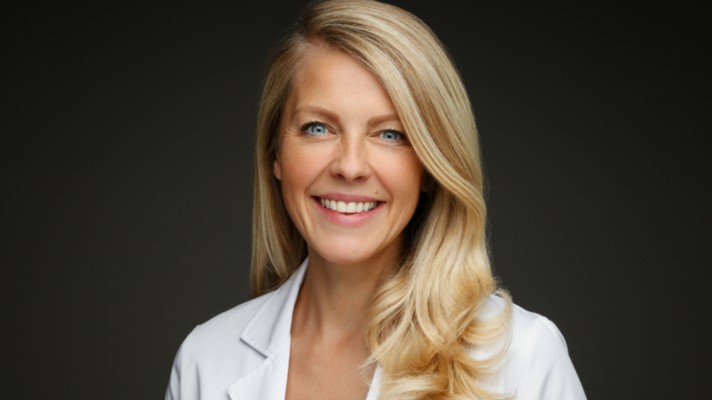 Dr Helen O' Neill from the shoulders up. She has blue eyes, long blonde hair and is smiling at the camera. She is wearing a white lab coat and the background is black.