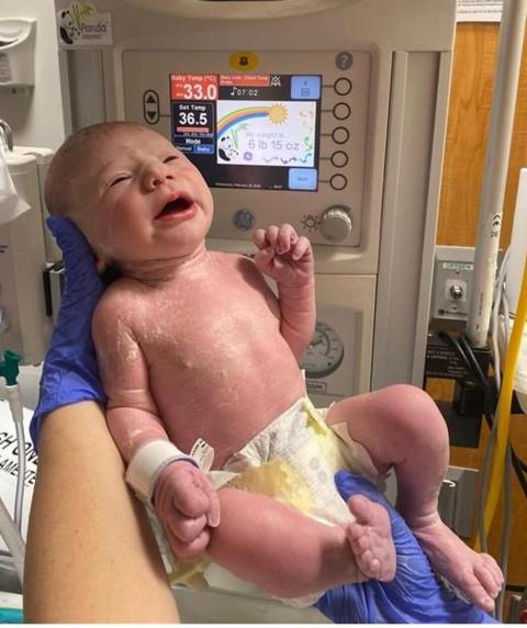 Newborn baby in the foreground, wearing only a disposable nappy and supported by gloved hands in front of a neonatal warming unit. A hospital identification band is attached to the baby’s wrist. The warming unit’s display screen shows temperature settings and a weight reading, with control buttons and gauges visible around it. Medical equipment, tubing and wall-mounted hospital fittings appear in the background. The image is taken in a clinical hospital setting.