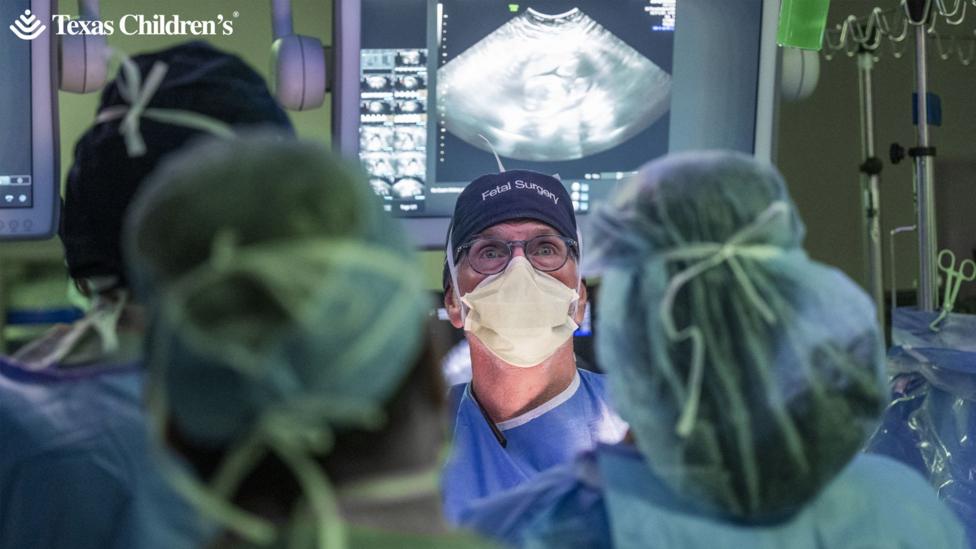 Medical staff in the foreground wearing surgical gowns, face masks and hair covers, gathered around an operating table. A person at the centre wears glasses, a face mask and a surgical cap labelled “Fetal Surgery”. Behind the team, large medical monitors display ultrasound images and diagnostic data. IV poles with hanging fluid bags and surgical instruments are visible at the sides of the operating theatre. The text “Texas Children’s” appears in the top-left corner of the image.