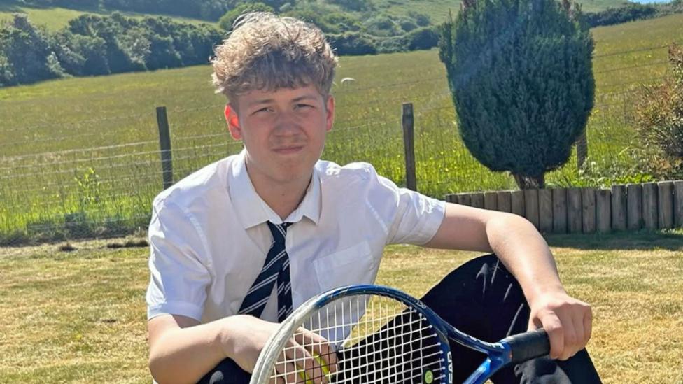 Jack sits in a garden that backs onto a large field in the countryside. He's wearing a school shirt and tie and is holding a tennis racquet.