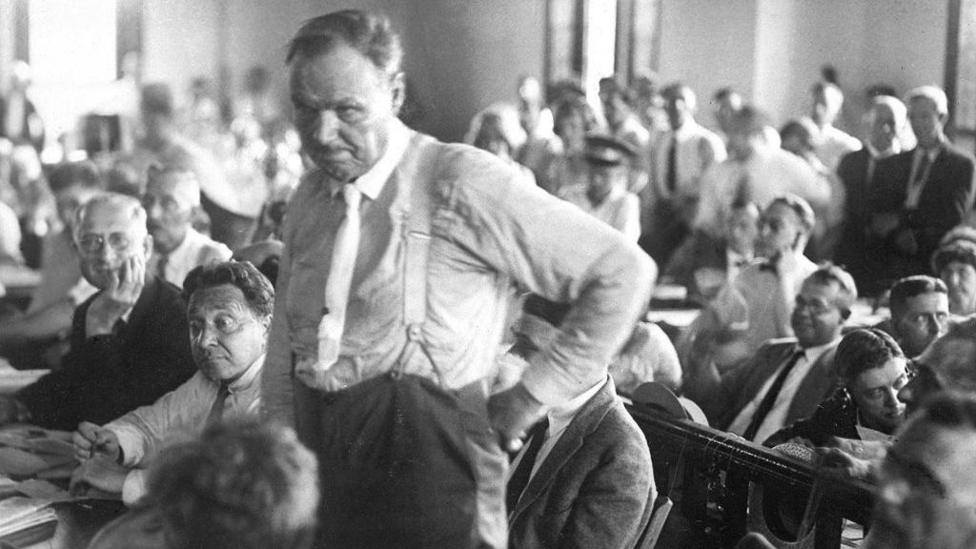 Chicago trial lawter Clarence Darrow standing at the front of a court listening to a judge. It's an old black and white photo
