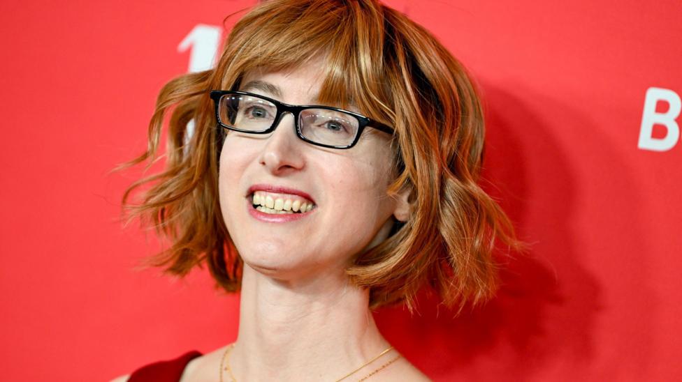 A woman wearing a red dress standing in front of a red backdrop. She is wearing a necklace, glasses and has short wavy hair