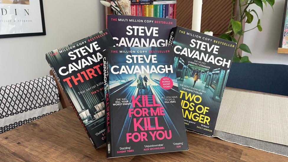 A handful of books from Steve Cavanagh sitting on a table in front of a book case.