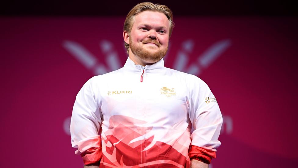 Mark Swan in red and white top standing on a podium, smiling.
