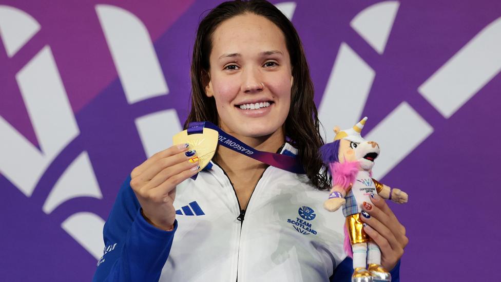Angharad Evans smiling on the podium, holding her gold medal and a little toy mascot.