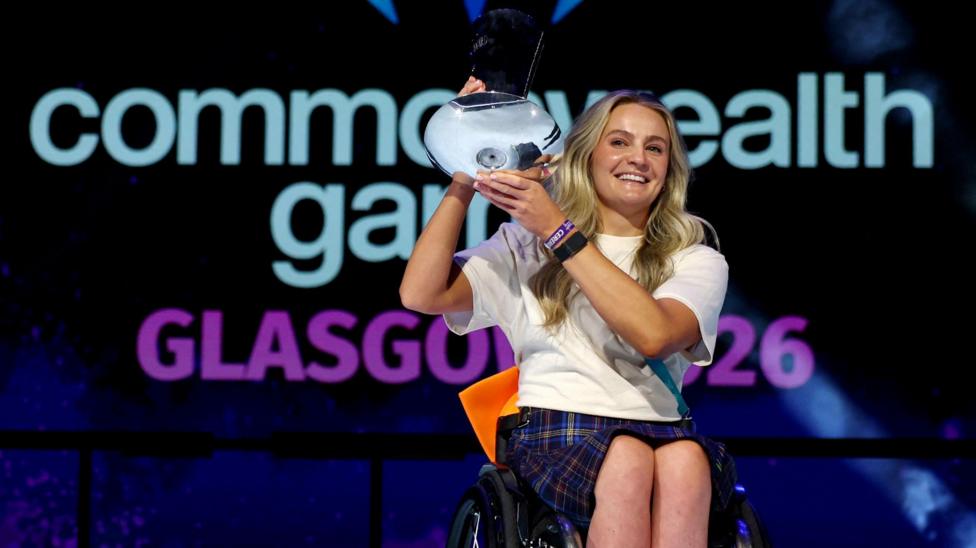 Melanie Woods holding the David Dixon Award in front of the Commonwealth Games logo. She is smiling broadly.