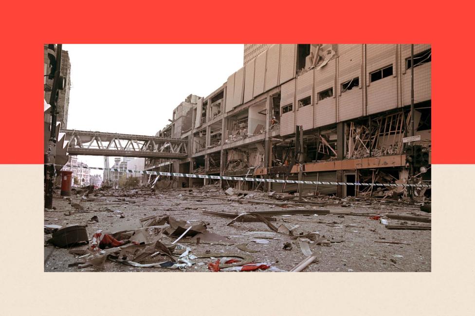 A photo dated 16 June 1996 shows the scene of devastation in Manchester City Centre following the bomb attack. Police tape stretches across the street and rubble is strewn over the road. Behind is the Arndale shopping centre. Its windows have been blown out.