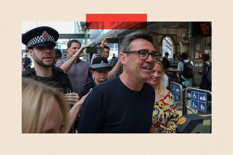 Newly elected Makerfield MP and former Greater Manchester Mayor Andy Burnham walks at Manchester Piccadilly station as he leaves for London, on 22 June 2026. He is a white man with dark greying hair and dark-framed glasses, and he wears a dark T-shirt. Behind him are two police officers. Two passers by are taking photographs of him with their phones.