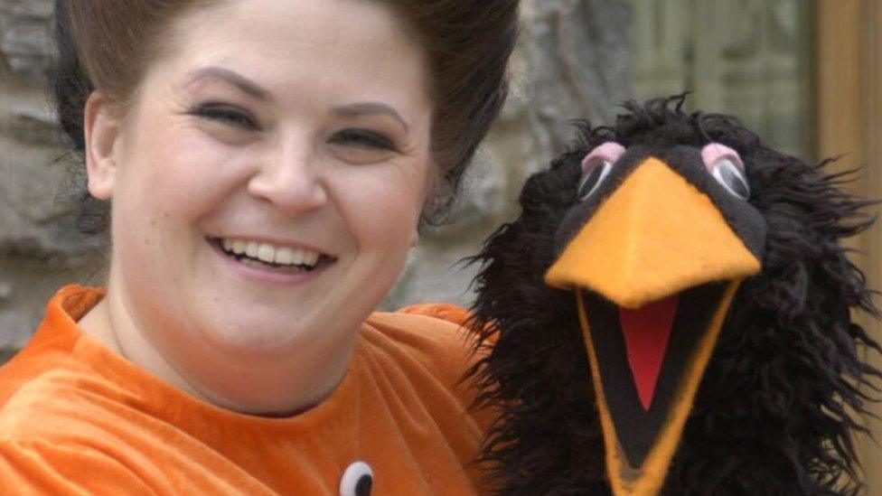 Rebecca Harries has brown hair which is tied back. She is wearing an oragne costume and holding a black bird puppet. She is smiling and looking at the camera.