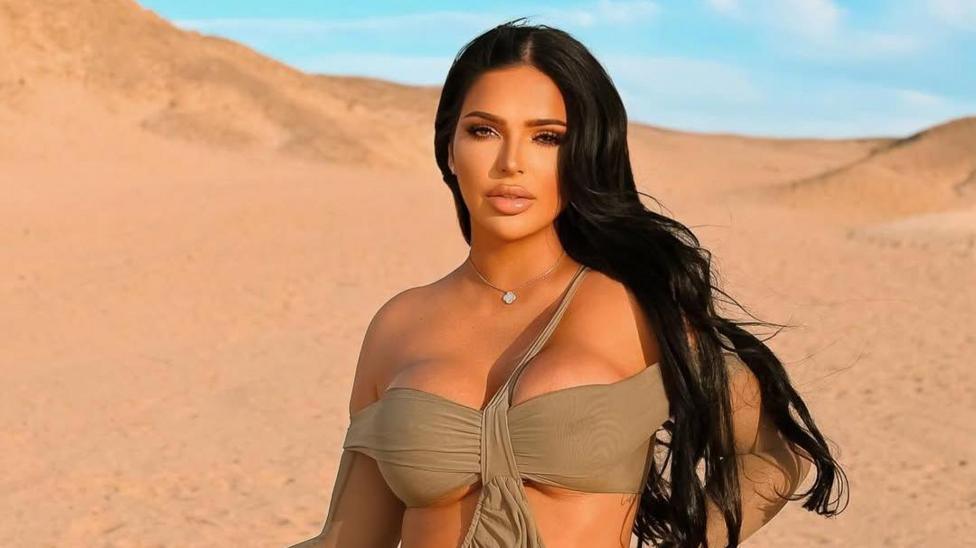 Klaudia Zakrzewska in a desert with blue sky. She wears an off-the-shoulder bikini style top which is taupe. Her hair is long and dark and behind her shoulders. She looks directly at the camera and has a small pendant necklace on. 