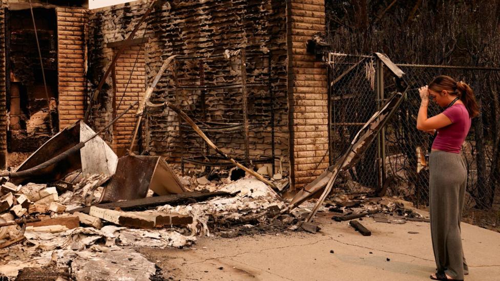 A woman holds her hand to her head as she looks at the rubble of a house. The bricks of the building are charred