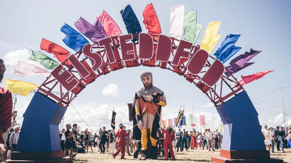 The puppet of Lord Rhys ap Gruffyd is guided through a giant blue arch, with Eisteddfod written in large letters upon it. There are different coloured flags flying from the arch and a crowd of people watching the procession.