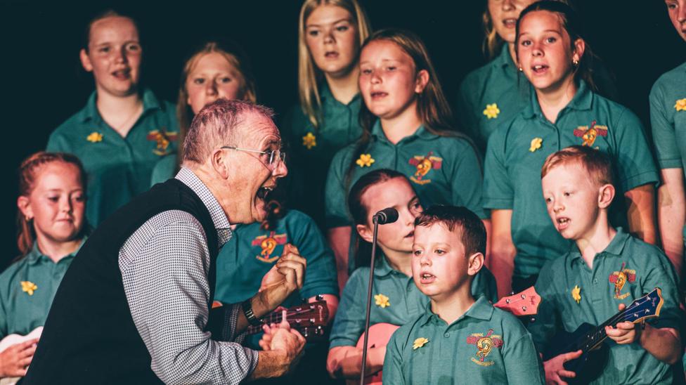A male conductor sings towards a group of young children in matching green school uniforms as they sing.