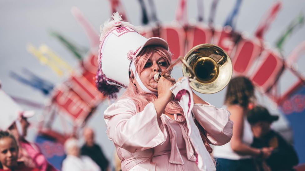 A woman wearing a pink costume and a pink parade hat blows into a trumpet.
