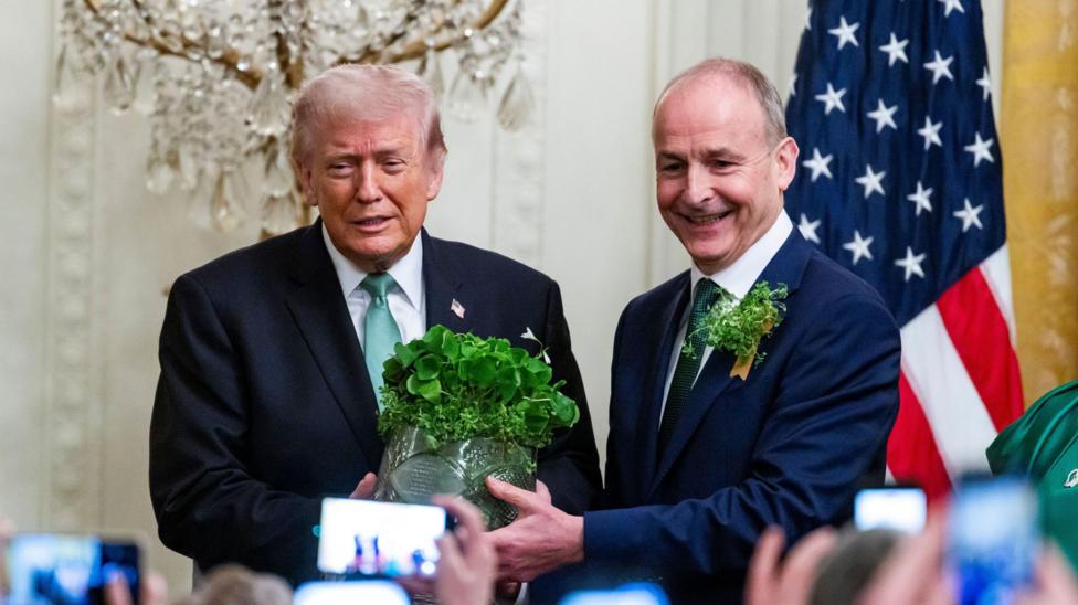 Donald Trump (left) beside Micheál Martin who is presenting him a shamrock bowl. Both wearing dark suits and green ties. Martin has a shamrock on his lapel. Both have grey hair.