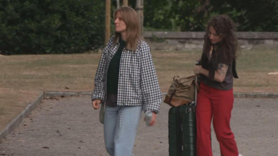 Image of Markéta Irglová with brown hair, and a black and white jacket. She's wearing blue jeans. Beside her is a woman with dark hair, black shirt and red trousers. She is carrying a suitcase.
