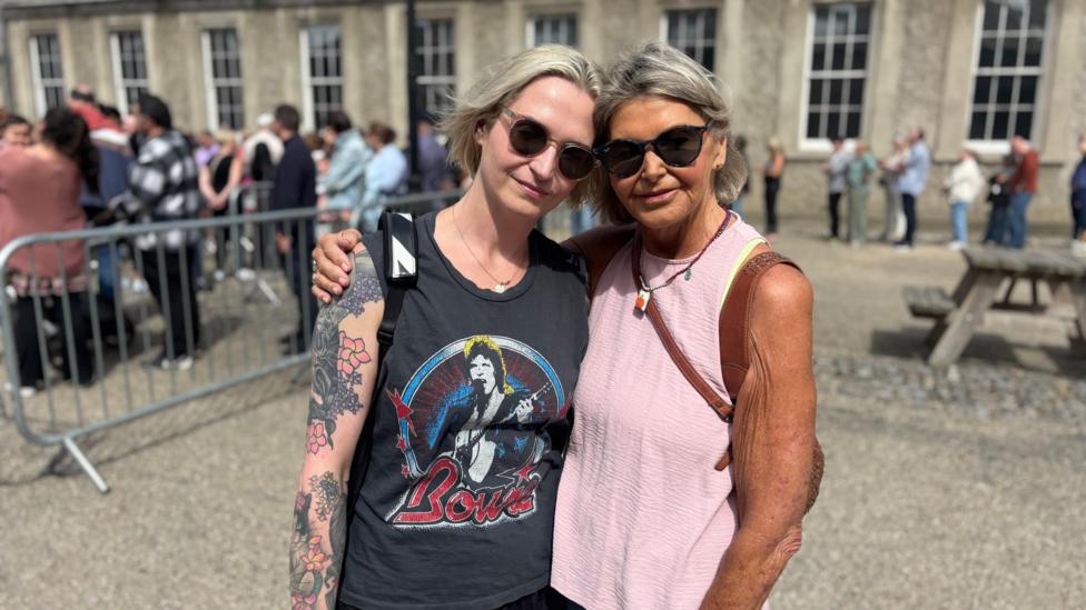 Two women pose for a photo, both are wearing best tops, on the left the woman has light hair and a navy top with a David Bowie design, on the right the woman has grey hair and is wearing a pink top.