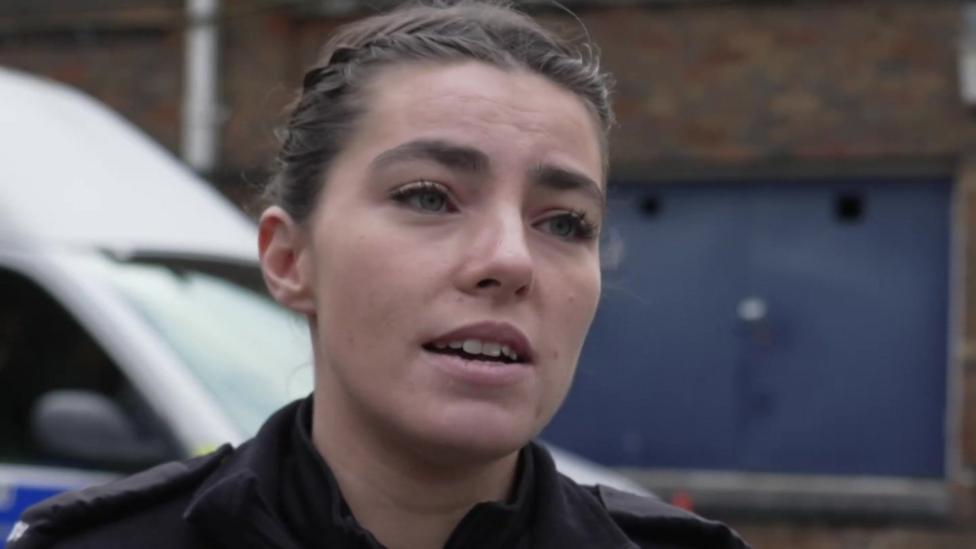 PC Jess Surman gives a TV interview outdoors. Her dark hair is tied back in braids and she wears a police uniform.