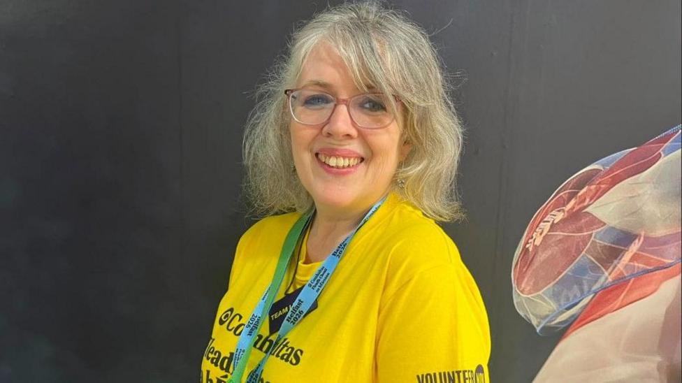 A woman, Camilla Reynolds pictured In front of a black wall with blue, white and red wall art on the right corner. She has grey hair and is wearing a yellow t-shirt with a blue lanyard and is wearing red glasses.