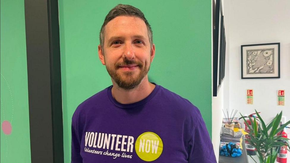A man, Toby Brusey pictured in an office. He is wearing a purple t-shirt that says 'Volunteer now, volunteers change lives' with a green circle graphic. behind him is a green and white wall with wall art and a plant.