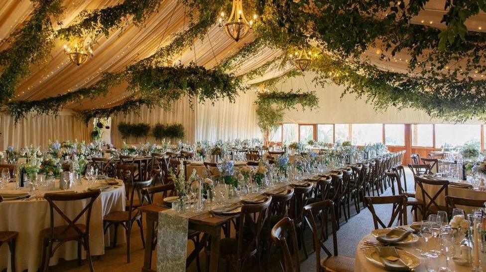 A wedding tent with green foliage and lights hanging from the ceiling. There are tables made up with flowers and cutlery ready for a wedding