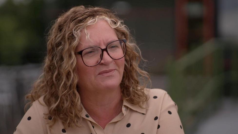 Catherine Marchant has shoulder length blonde wavy hair and black rimmed glassed. She wears a beige shirt with black polka dots. She looks deep in thought, away from the camera.