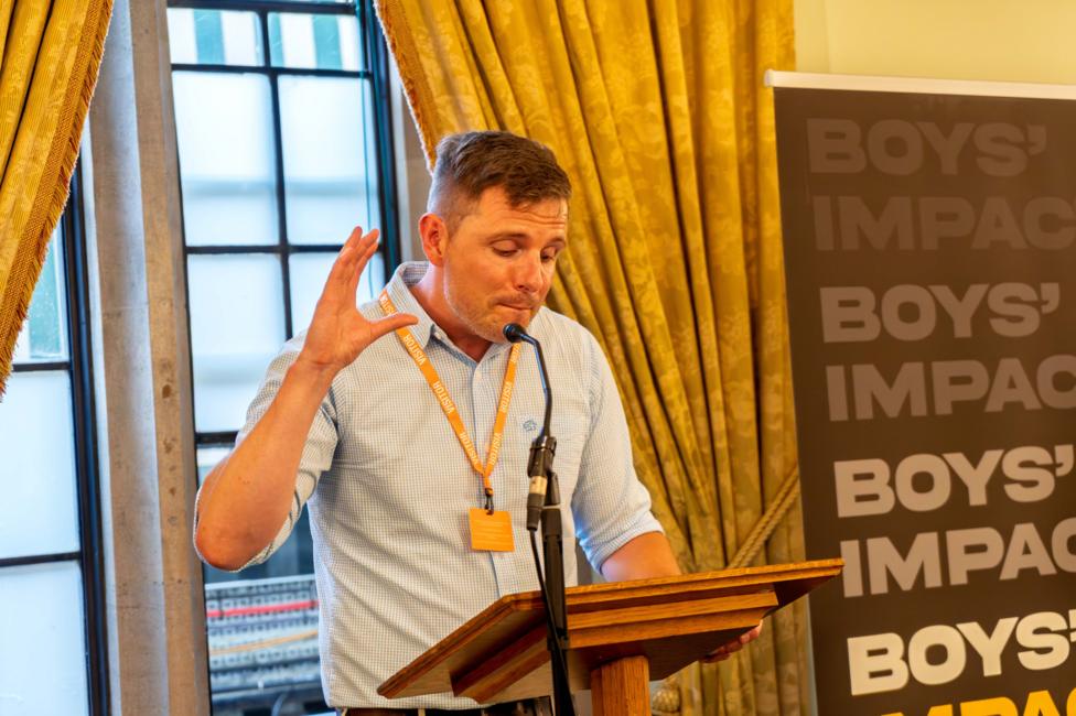 Dr Alex Blower is standing at a wooden lectern speaking into a microphone. He is wearing a blue checked shirt with his sleeves rolled up and is looking down at notes while he talks into the microphone. Behind him is a impact with the Boys' Impact logo.