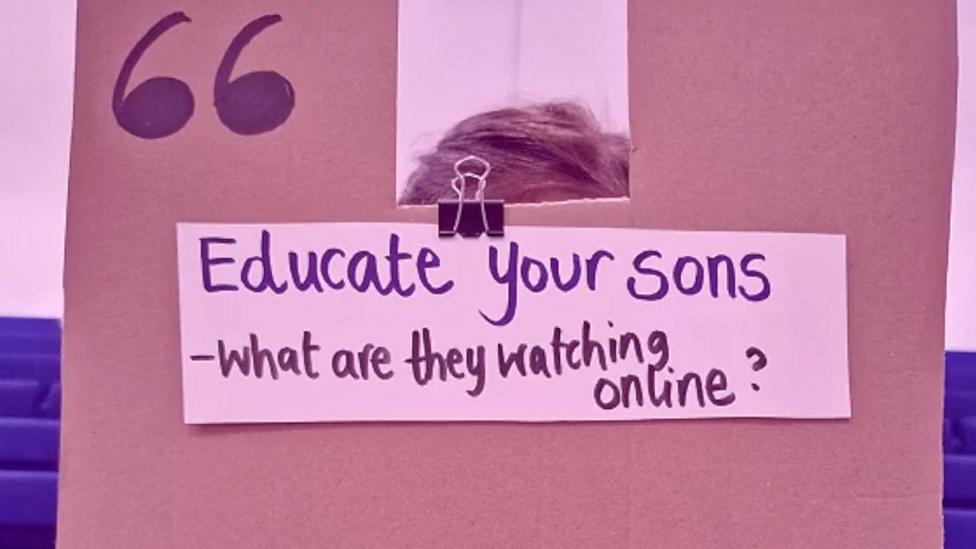 A cardboard sign is held in front of a girls face, her hands are visible on the edges. 'Educate your sons - what are they watching online?' is written in black marker pen.