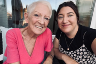 Two women smiling at the camera. One on the left has a pink top and short white hair, the other has dark hair and is wearing a black t-shirt under a flowery dress.