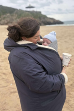 A woman on a beach in a blue coat. She has a baby tucked inside her coat and holds a coffee cup. She is on the beach.