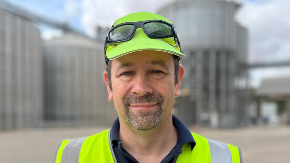 A man - Simon Willis - looking straight at the camera. He is standing in front of several silos and is wearing a yellow high visibility vest and matching hat.