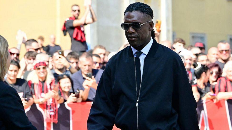 Clarence Seedorf, wearing sunglasses and a black jacket, walks with his head bowed