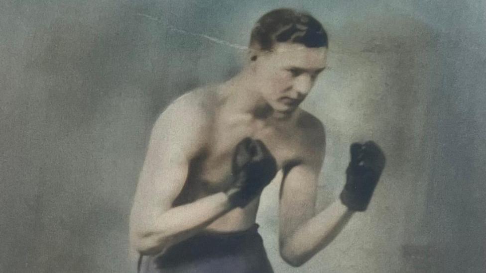 An old photograph of Maynard Powell wearing boxing gloves. He has short dark hair and is not wearing a shirt.
